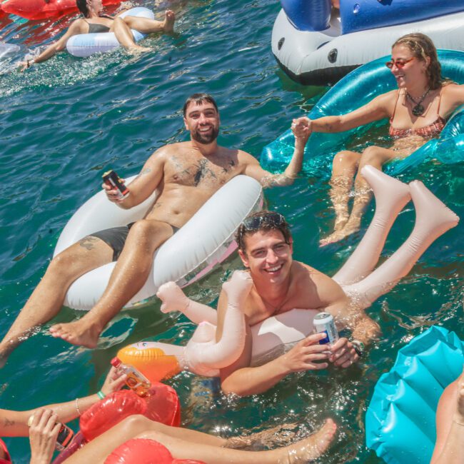 A group of people are relaxing and laughing on colorful pool floats in clear blue water, holding drinks and enjoying the sunshine at a lively outdoor gathering.