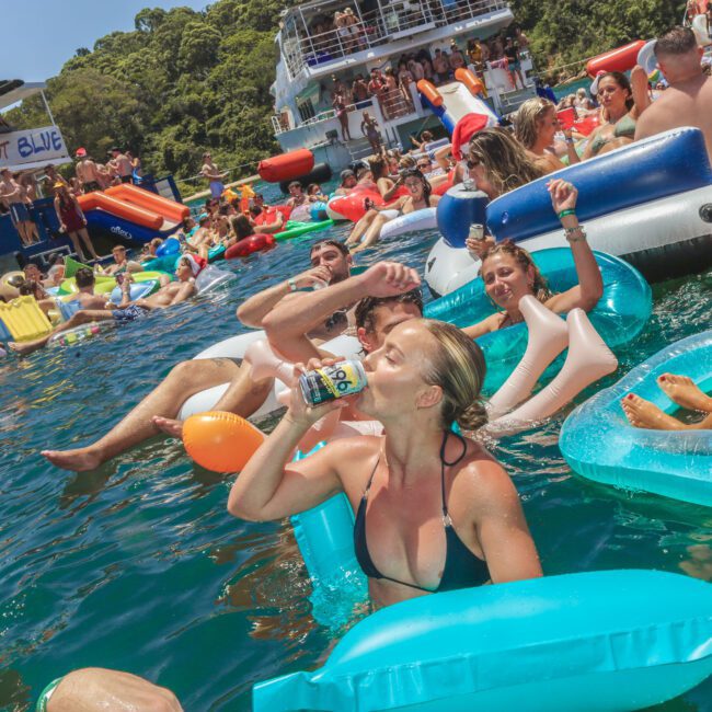 A large group of people lounge on colorful inflatables and swim in the water near boats during a lively summer party. A woman in front drinks from a can, and everyone appears to be enjoying the sunny, festive atmosphere.