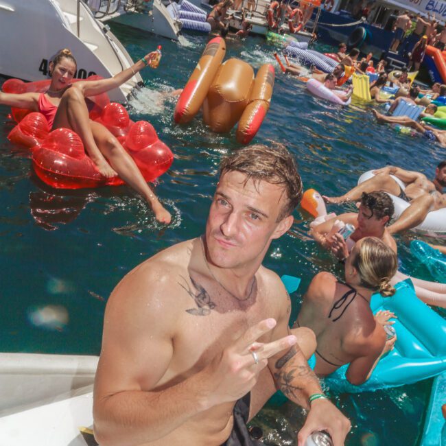 A man in swim trunks sits on a boat step, holding a drink and making a peace sign. Behind him, people relax on colorful floats and pool toys in the water at a lively party with boats and sunshine.