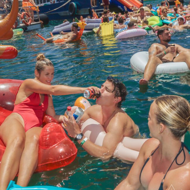Group of people relaxing on colorful pool floats in clear blue water, with a boat and more partygoers in the background. A woman in a red swimsuit hands a drink to a man on a float, while others laugh nearby.