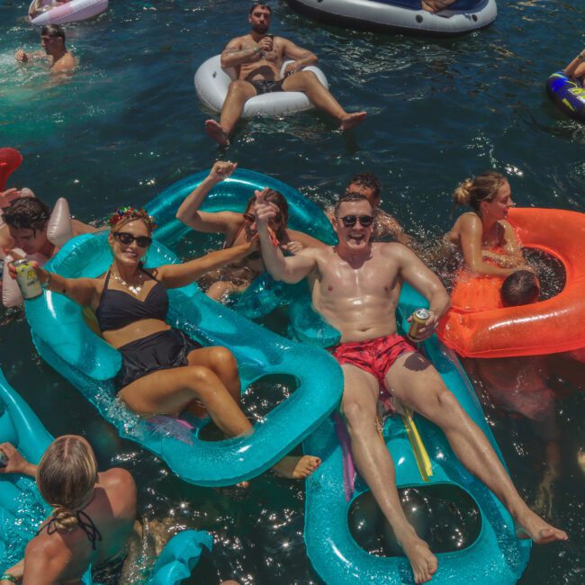 A group of people relax and have fun on colorful inflatable floats in a crowded, sunlit body of water, raising drinks and smiling during a summer party. The atmosphere is lively and joyful.