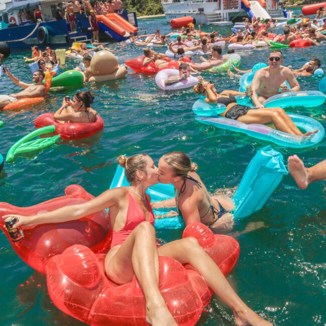 A group of people relax and party on colorful pool floats in the water near boats. Two women in swimsuits kiss on a red bear-shaped float, surrounded by others enjoying the sunny day.