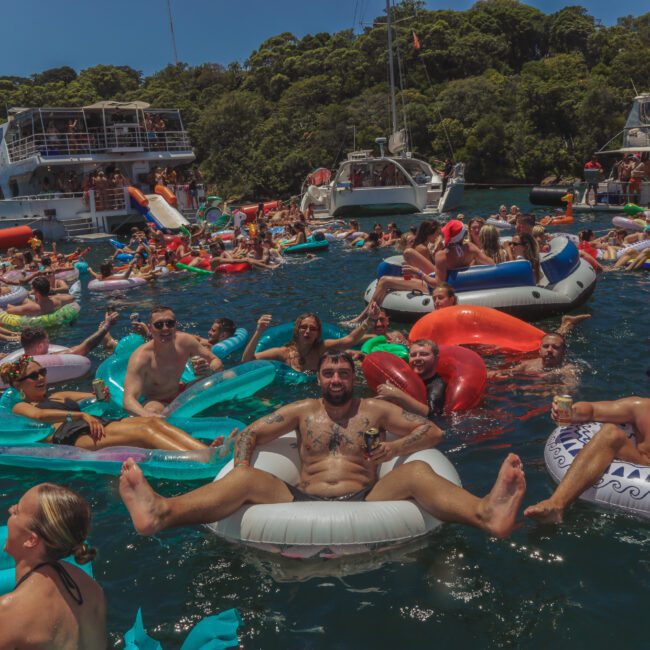 Dozens of people relax on colorful inflatables in the water near several boats, surrounded by lush green trees on a sunny day. Many are smiling and enjoying a lively, festive atmosphere.
