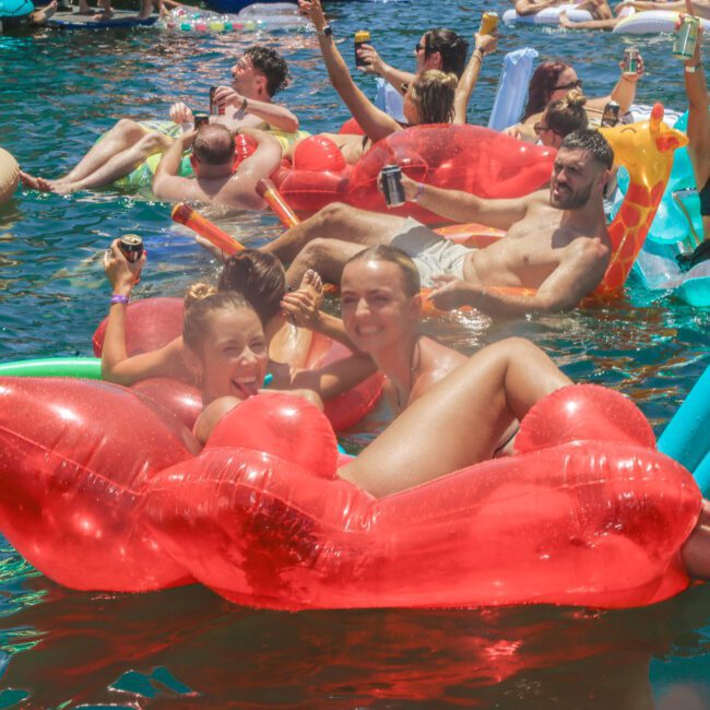 People relax and smile on colorful inflatable floats in a crowded pool, enjoying a sunny day. Two women are in the foreground on a red float, holding drinks and laughing, surrounded by others on various inflatables.