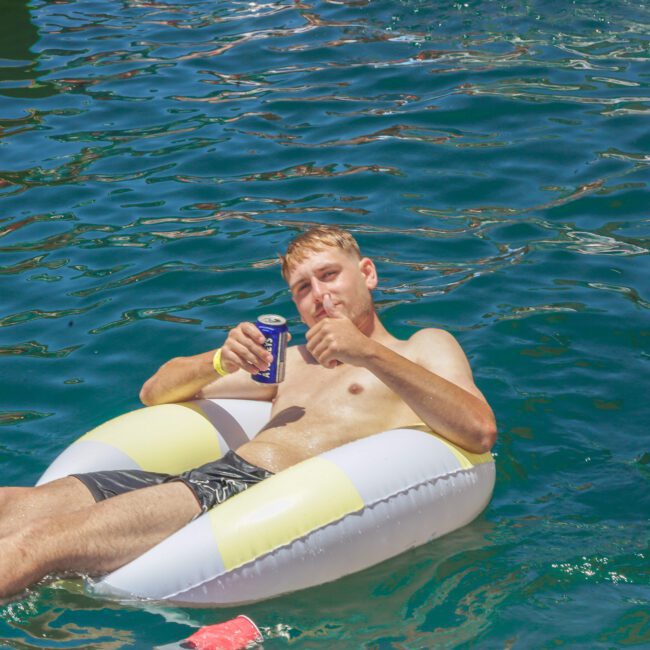 A man relaxes on a white and yellow inflatable pool float in the water, holding a can and giving a thumbs up, with sunlight reflecting on the surface around him.