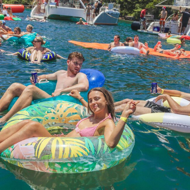 People relax on colorful pool floats in a lake near boats, holding drinks and enjoying sunny weather. Others swim and lounge in the background at a lively outdoor party. The event logo "Yacht Social Club" is visible.