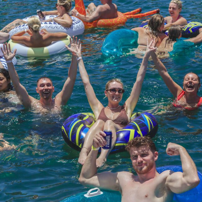 A group of people having fun in the water with colorful pool floats. Some are smiling with raised arms, one person flexes their arm, and others relax in the background. The scene is lively and joyful.