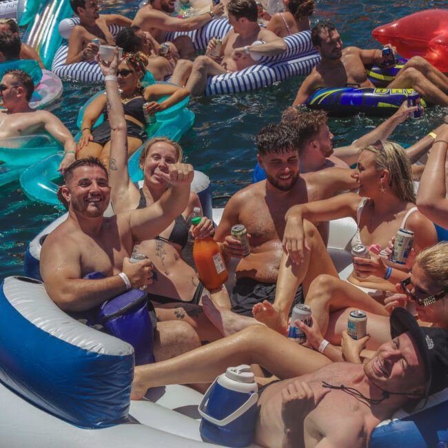 A group of young adults relax on pool floats, laughing and holding drinks, surrounded by others on inflatables in bright blue water at a lively outdoor party. The mood is festive and joyful.