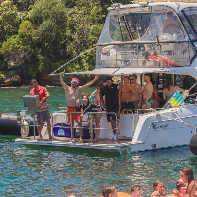 A group of people stand and pose on a white boat docked on clear water, surrounded by lush greenery. Some are smiling and waving, while others relax. People in life jackets are seen swimming nearby.