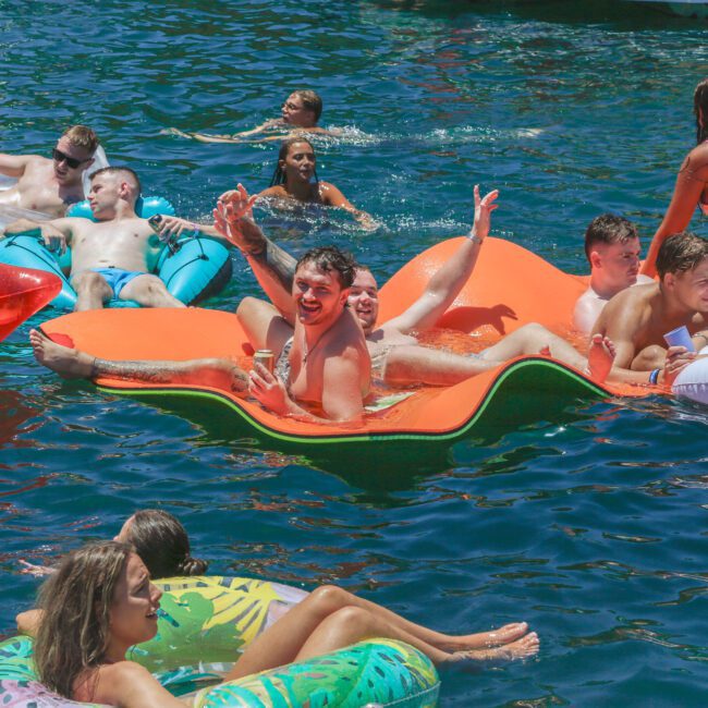 A group of young adults relax and have fun on colorful inflatable floats in a sunny lake, laughing and enjoying the water together.