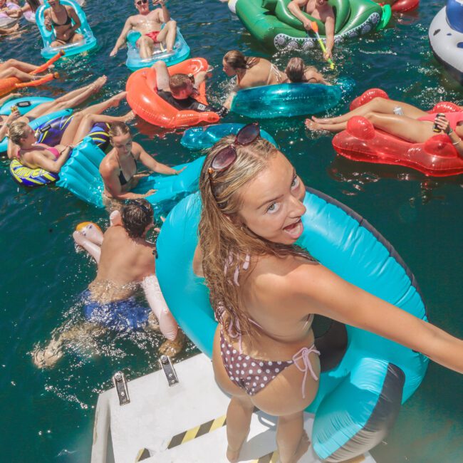 A woman in a polka dot bikini holds a blue inflatable ring and smiles at the camera while standing on a boat ladder. Around her, many people relax and float on colorful inflatables in the water under bright sunlight.