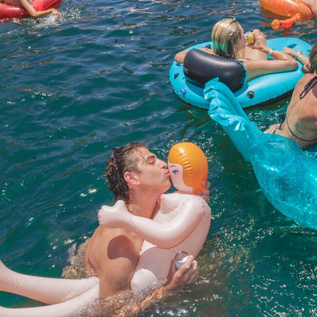 A man in a pool kisses an inflatable doll while holding a drink. Around him, other people lounge and float on colorful pool inflatables, enjoying a sunny day in the water.