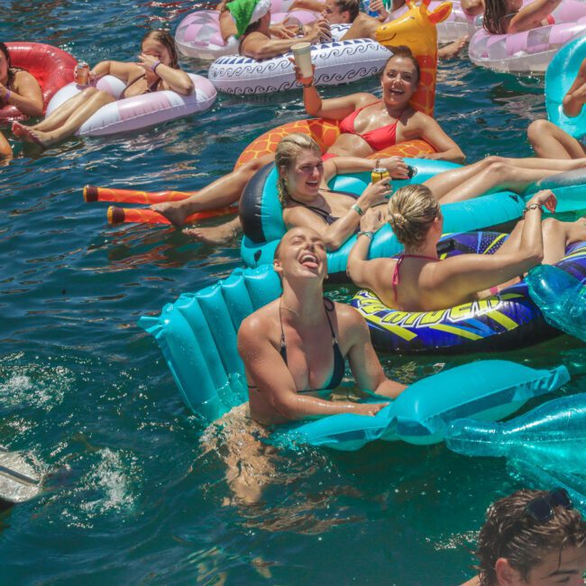 A group of people float and relax on colorful inflatable rafts in a sunny, crowded swimming area, laughing and enjoying drinks together on the clear blue water.
