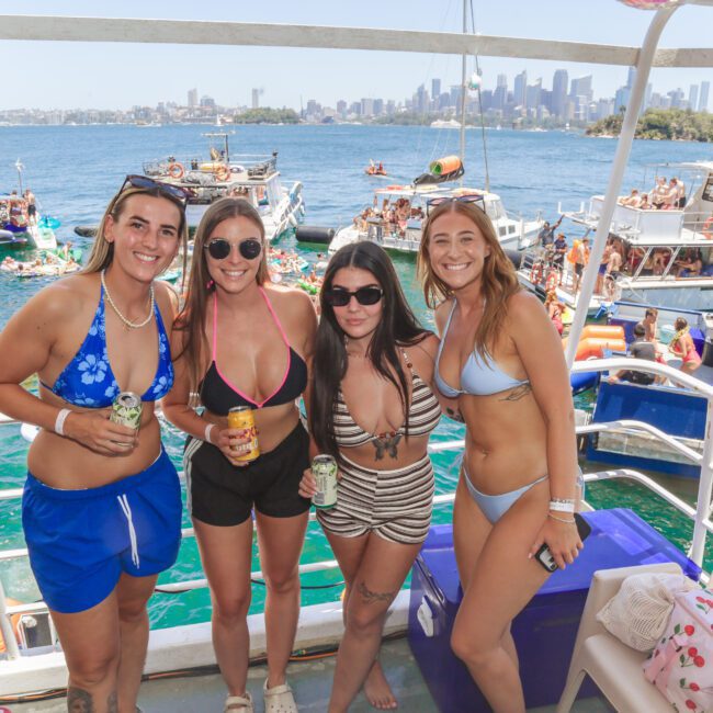 Four women in swimsuits stand smiling on a boat with drinks in hand. In the background, people on boats and inflatables enjoy a sunny day on the water, with a city skyline visible in the distance.