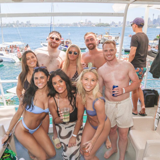 A group of young adults in swimsuits smile and pose together on a boat, holding drinks. The background shows water, other boats, and a city skyline under a clear, sunny sky.