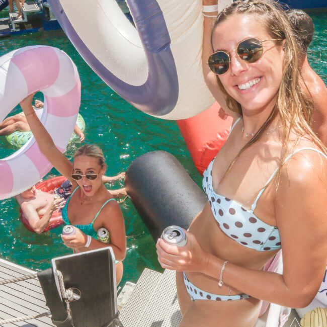 Two women in swimsuits, holding drinks and large inflatable floaties, smile by the water at a dock. Other people and colorful floats are in the background, suggesting a lively summer pool or boat party.