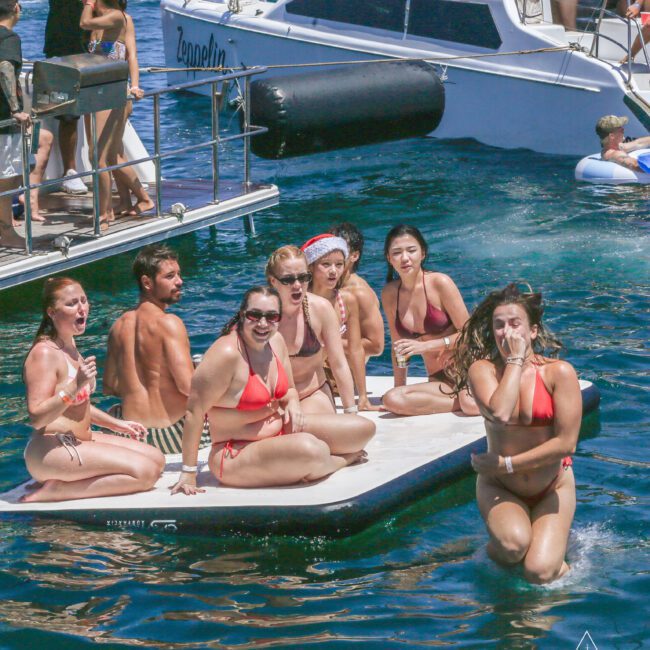 A group of people in swimsuits sit and pose on a floating platform in the water near a yacht, with others in the background enjoying the sun. Some are smiling and laughing, and one person wears a Santa hat. The scene is lively and festive.