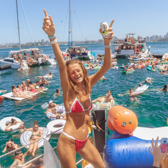 A woman in a red bikini smiles and raises her arms while standing on a boat at a lively pool party, surrounded by people on boats and inflatables in bright blue water with a city skyline in the background.