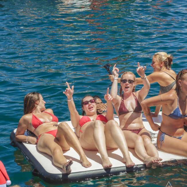 A group of women in swimsuits relax and laugh together on a floating mat in the water, surrounded by boats and sunshine, enjoying a lively day outdoors.