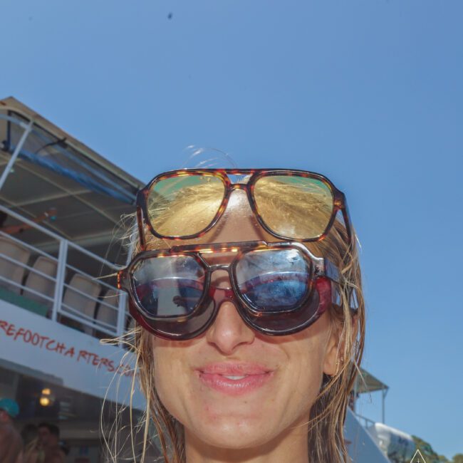 A woman smiles under a clear blue sky, wearing three pairs of sunglasses stacked on her face. People and a boat are visible in the sunny background, with a "Yacht Social" logo in the corner.
