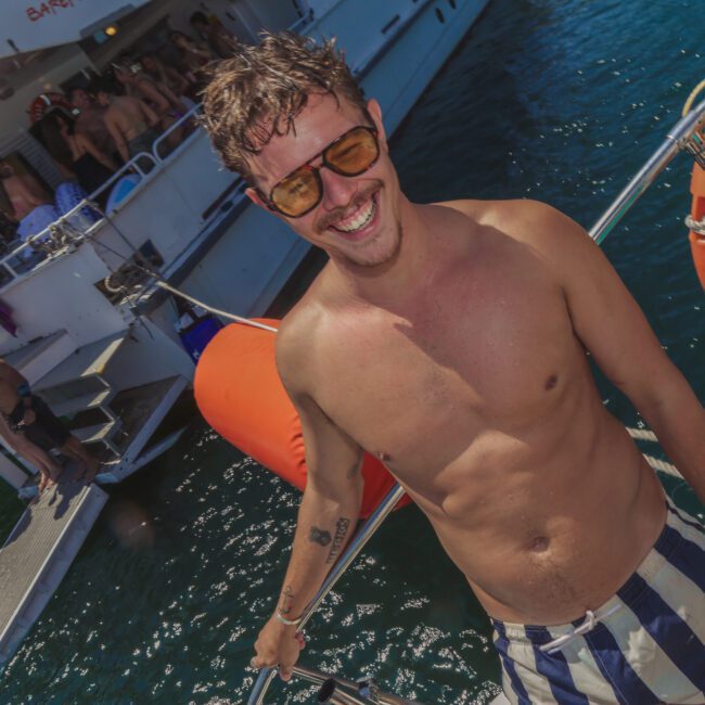 A smiling shirtless man wearing sunglasses stands on a boat, with people partying on another boat in the background. The scene is sunny, festive, and set on clear blue water.