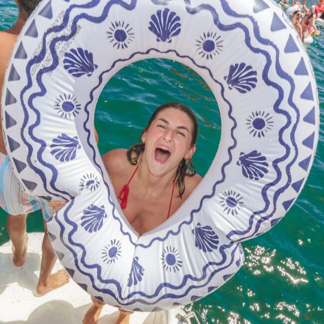 A young woman in a swimsuit smiles and laughs through a decorative inflatable ring on a boat, with clear blue water and other people swimming and floating nearby. The scene is lively and sunlit.