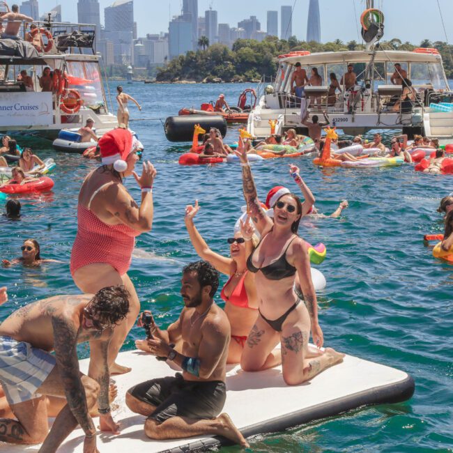 People in swimsuits celebrate on the water with inflatables and boats, some wearing Santa hats. The city skyline and a bridge are visible in the background under sunny skies. The scene is lively and festive.
