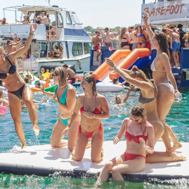 A group of young women in swimsuits laugh and pose on a floating platform in the water, surrounded by people swimming and boats with slides in a lively summer party atmosphere.