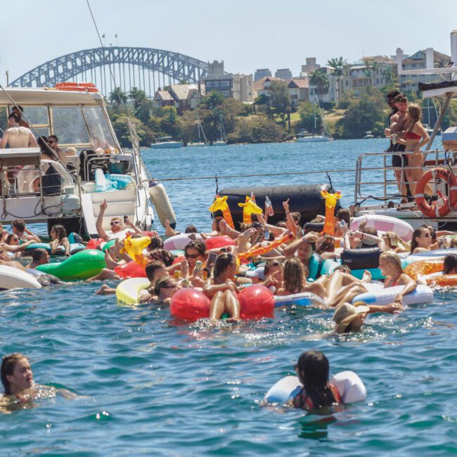 A large group of people relax on colorful inflatable pool floats in the water near boats, with the Sydney Harbour Bridge visible in the background on a sunny day.
