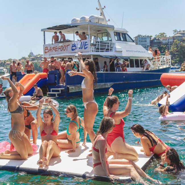 A group of people in swimsuits dance and relax on a floating platform in the water, with party boats, slides, and more people in the background on a sunny day.