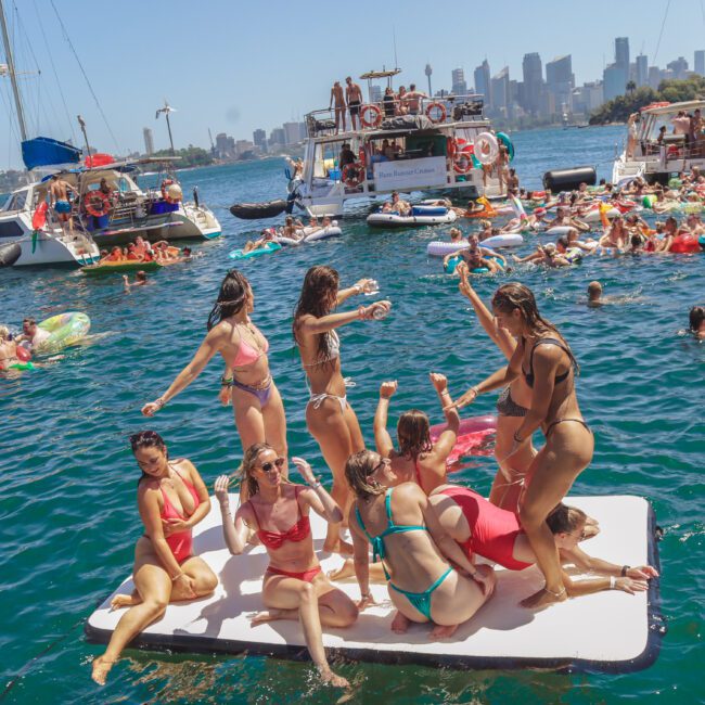 A group of people in swimsuits gather on a floating platform in the water, surrounded by boats and others swimming or lounging on inflatables, with a city skyline in the background.