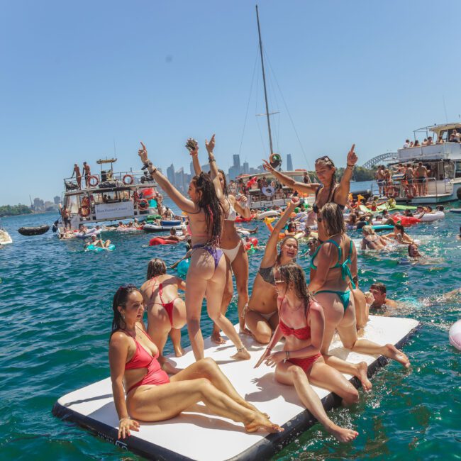 A group of people in swimsuits dance and pose on a floating platform surrounded by others swimming, floating on inflatables, and boating at a lively water party under a sunny, clear sky.