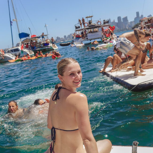 A woman in a bikini sits on the edge of a boat, smiling at the camera. Behind her, people swim and play on a floating mat, with several boats and a city skyline in the background under a clear sky.