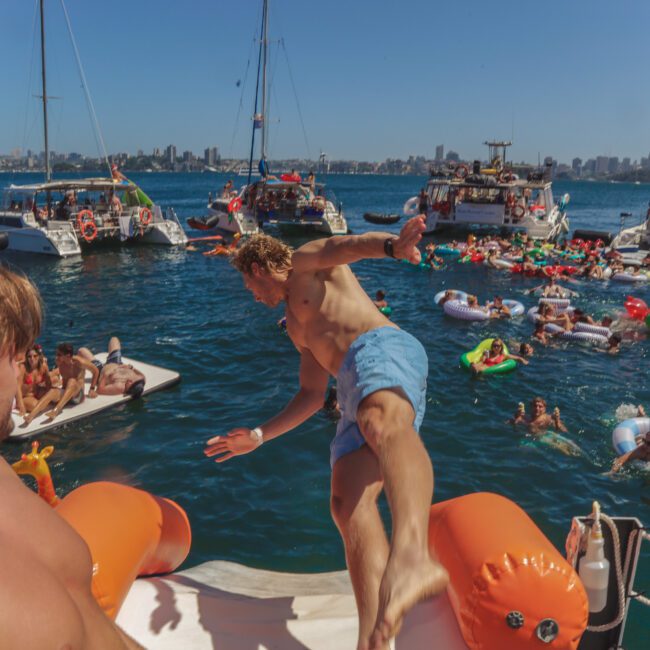 A shirtless man in blue swim trunks jumps off a boat into crowded water filled with people on inflatables and boats, with a sunny city skyline in the background.