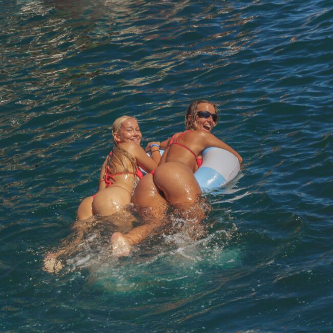 Two women in swimsuits smile as they lay on an inflatable float in bright blue water. Sunlight reflects off the waves. The scene appears joyful and summery. “Yacht Social Club” is written in the corner.