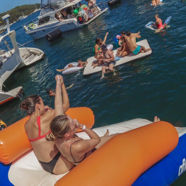 People relax and have fun on inflatable floats and boats in the water. Two women in swimsuits sit on a large float in the foreground, while more people socialize on nearby platforms and boats in the sunny scene.