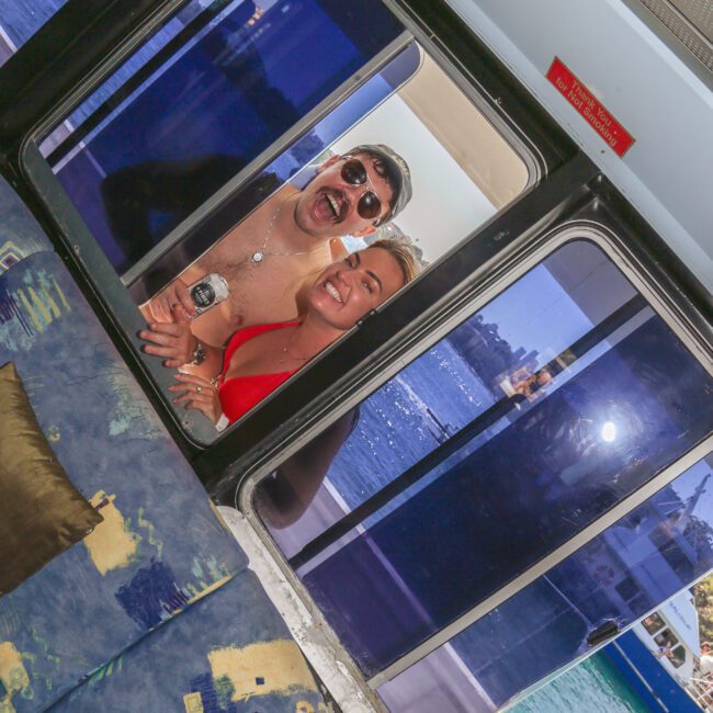 Two people in swimsuits smile and pose playfully outside a glass door on a boat, with blue water and city skyline visible in the background. The photo is taken from inside, showing part of a patterned couch.