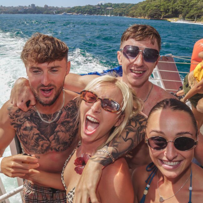 A group of young adults in swimsuits smile and pose together on a boat, with blue ocean water and a forested coastline in the background under a sunny sky.