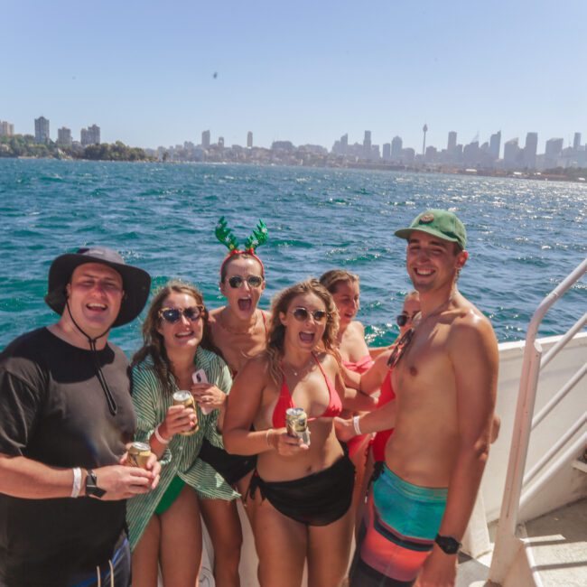 A group of six young adults in swimwear smiling and holding drinks on a boat, with a city skyline, blue water, and clear sky in the background. One person wears green antenna headbands.