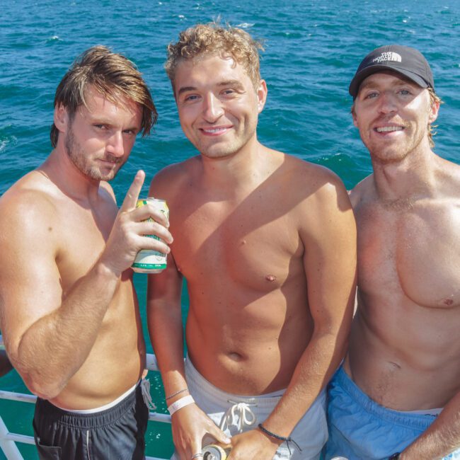 Three shirtless men stand close together on a boat, smiling at the camera with the ocean in the background. One man holds a drink, and it is sunny and bright.