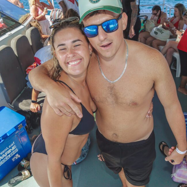 A young woman and man in swimsuits smile at the camera on a boat. The man wears sunglasses and a cap, with his arm around the woman. Other people sit and relax in the background. The scene is sunny and casual.