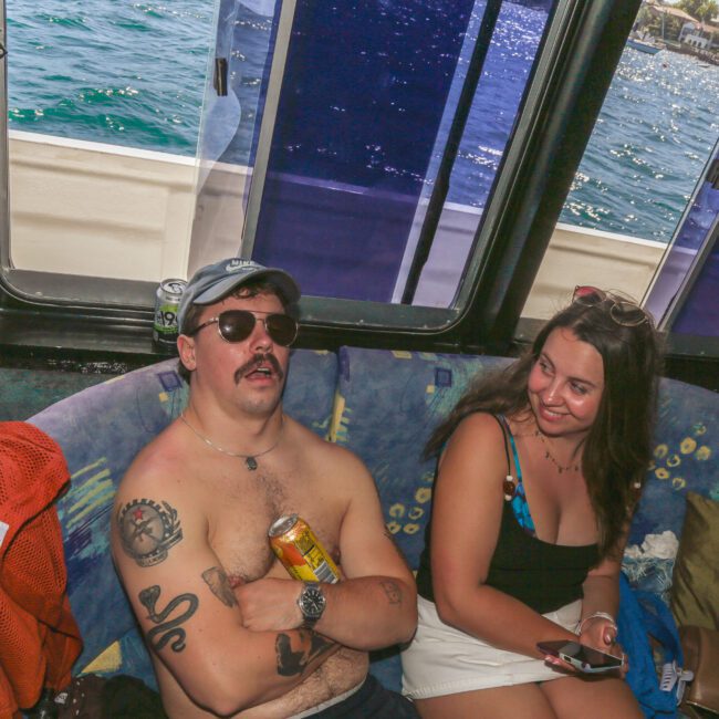 A shirtless man wearing sunglasses and a cap relaxes on a boat sofa holding a drink, while a smiling woman in a swimsuit sits beside him. Sunlight streams in and the ocean is visible through the windows behind them.