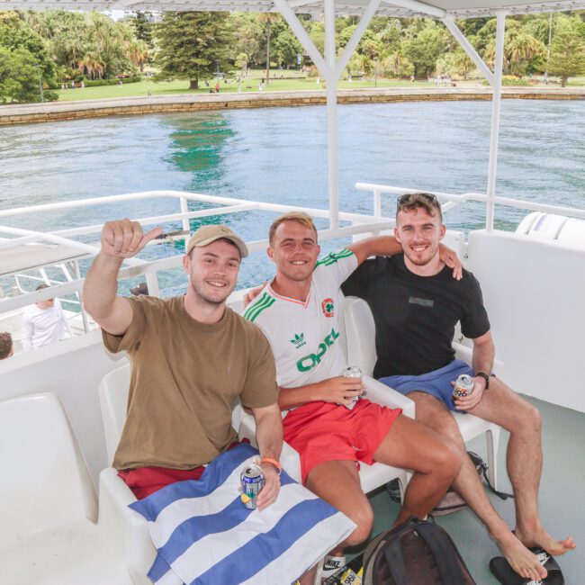 Three smiling men sit on a boat deck, holding drinks and posing for a photo. They are dressed in casual summer clothes, with greenery and water visible in the background on a sunny day.