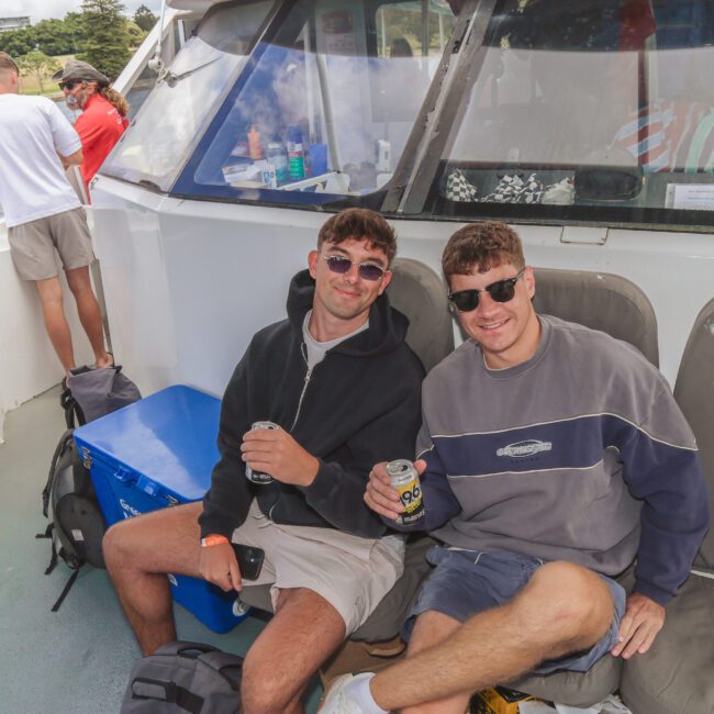 Two men wearing sunglasses and casual clothes sit on a boat, smiling and holding drinks. Other people stand nearby, and water with trees is visible in the background. The "Yacht Social Club" logo is in the corner.