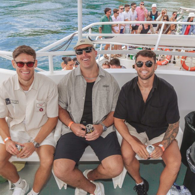 Three men in casual summer clothing and sunglasses sit smiling on a boat, each holding a drink. Behind them, other people socialize on a lower deck over water on a sunny day.