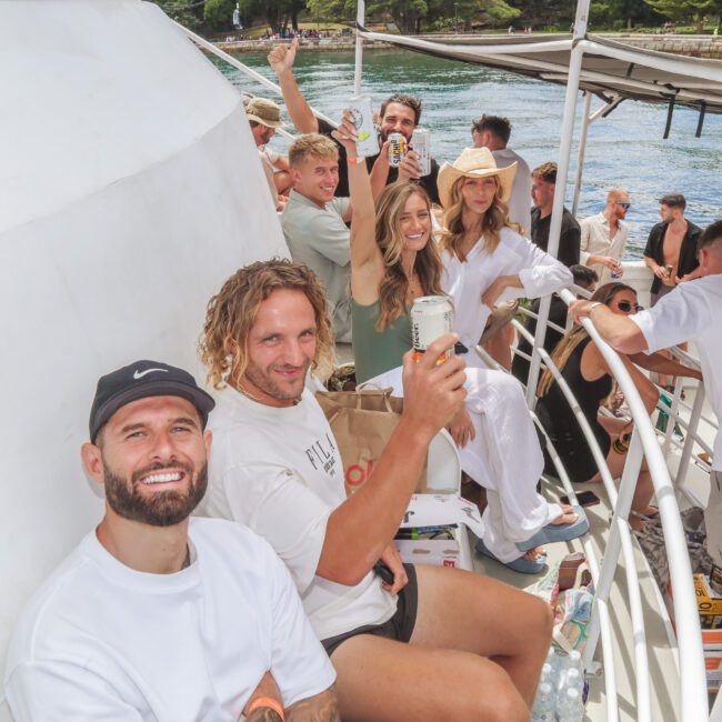 A group of people smile and enjoy drinks together on a boat under sunny weather, with greenery and water in the background. Some are seated, while others stand, all looking cheerful and relaxed.
