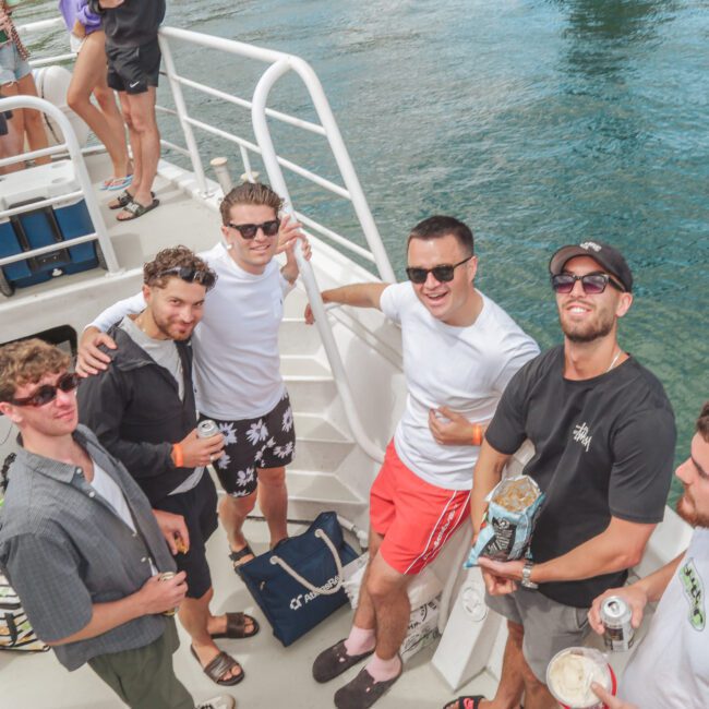 A group of six young men, casually dressed and smiling, stand together on the deck of a boat holding drinks. The boat is on clear blue water and other people can be seen in the background.