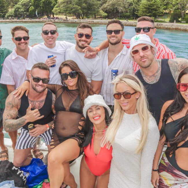 A group of men and women in swimsuits and summer clothes pose and smile together on a boat with clear blue water and green trees in the background. Some are holding drinks and wearing sunglasses.