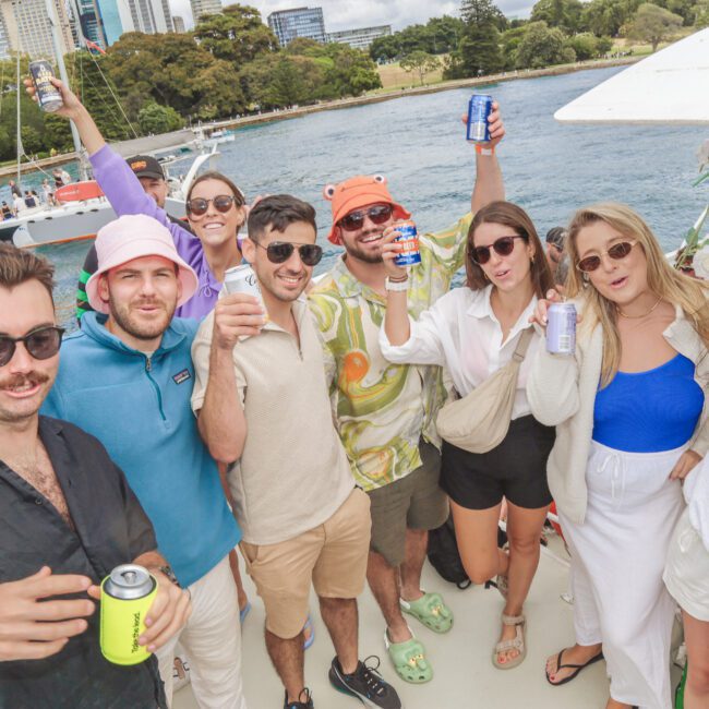 A group of eight friends stand on a boat, smiling and holding up drinks. They are wearing summer clothing and sunglasses. Water and trees are in the background, along with tall buildings. It’s a sunny day.