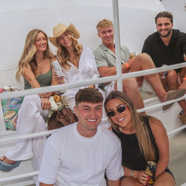 A group of six young adults smiling and sitting together on a boat deck, holding drinks. Some are wearing casual summer clothes and hats. The atmosphere appears relaxed and cheerful.